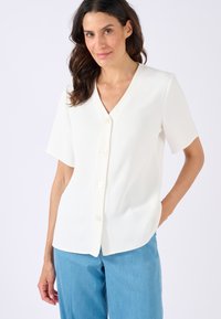 Blusa bianca a maniche corte con scollo a V, chiusura anteriore con bottoni, texture morbida e orlo arrotondato. Indossata con pantaloni azzurri chiari.