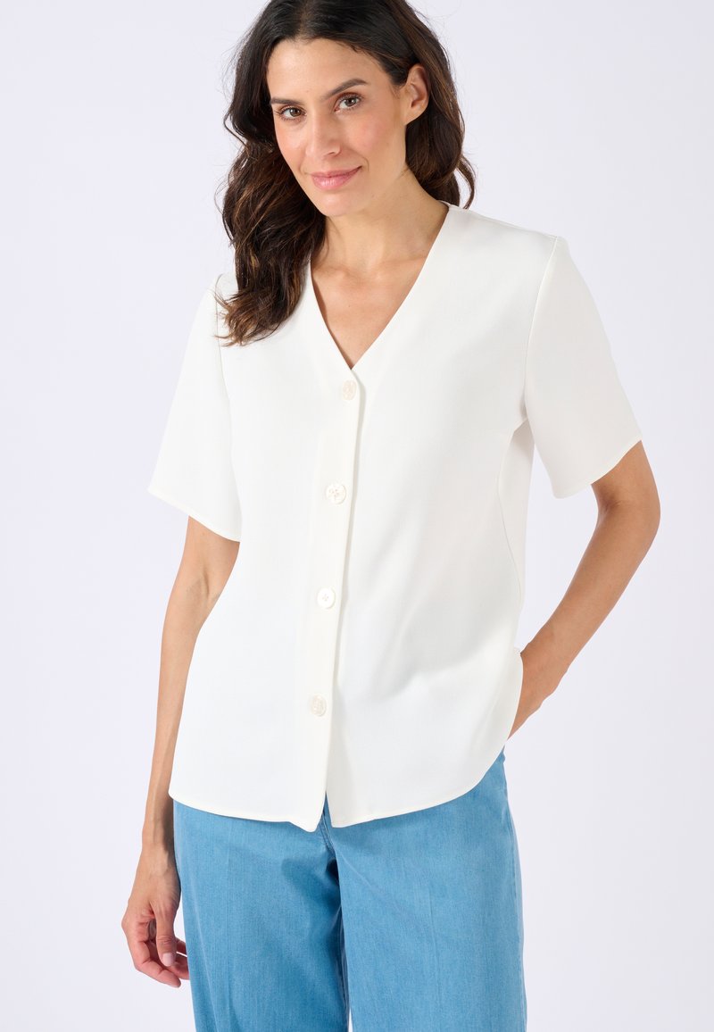 Blusa bianca a maniche corte con scollo a V, chiusura anteriore con bottoni, texture morbida e orlo arrotondato. Indossata con pantaloni azzurri chiari.