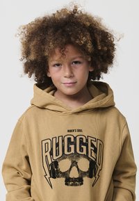 Dieťa s kučeravými vlasmi, oblečené v béžovej mikine s kapucňou, na ktorej je nápis "RUGGED" a grafika lebky s slúchadlami, na čistom bielom pozadí.