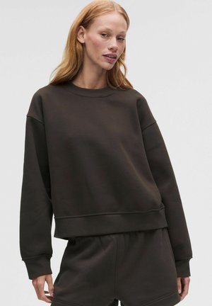 Frau mit roten Haaren trägt einen lockeren schwarzen Pullover und passende Hose und steht vor einem schlichten hellen Hintergrund.