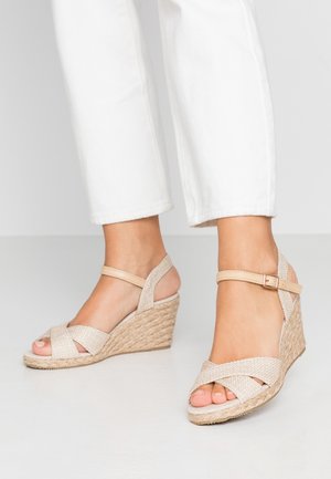 Fødder iført beige flettede kilehæls-sandaler med anklerem kombineret med hvide cropped bukser på en ensfarvet lys baggrund.