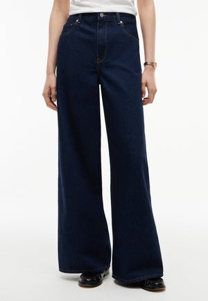 Person iført mørkeblå vidde jeans, hvid skjorte, guld armbåndsur og sorte sko på en hvid baggrund.