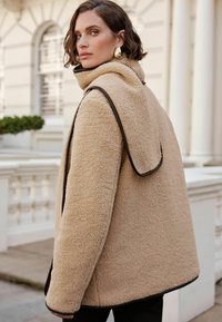 Beige shearlingjacka med hög krage och sidoflappar. Brunt kantband längs kanterna. Avslappnad passform, långa ärmar och synlig sömdetalj.