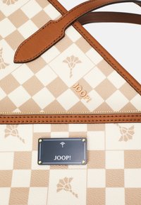 Béžová a bílá kostičkovaná kabelka s hnědými koženými rukojetmi a ozdobnými prvky, s květinovými motivy a černou značkovou destičkou s nápisem "JOOP!"