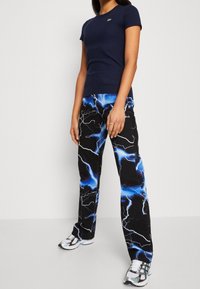 Žena v námořnickém modrém tričku s krátkým rukávem, černých kalhotách s modro-bílými vzory blesků a stříbrno-bílých sportovních botách.