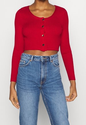 Kvinde iført en rød ribbet cropped sweater med hjerteformede knapper og højttaljede blå jeans, stående mod en ensfarvet baggrund.