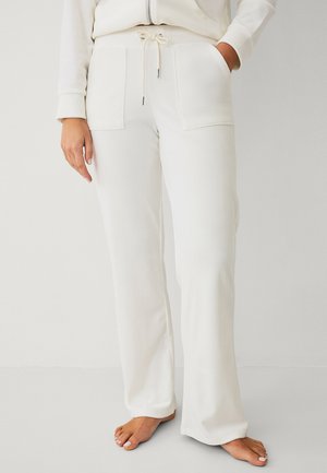 Osoba nosząca białe, szerokie spodnie dresowe z kieszeniami na przodzie i paskiem ściągającym w talii, stojąca boso na jednolitym tle.