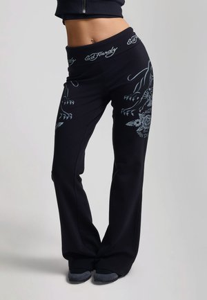 Mujer que lleva pantalones negros Ed Hardy con diseños florales grises y garras de tigre, combinados con zapatos negros y un top negro recortado.