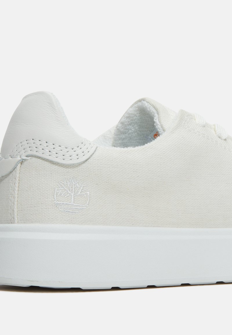 Zapatillas de lona blancas con un detalle de talón de cuero blanco, que presentan un sutil logo en relieve, costuras tonal y una suela de goma texturizada blanca.