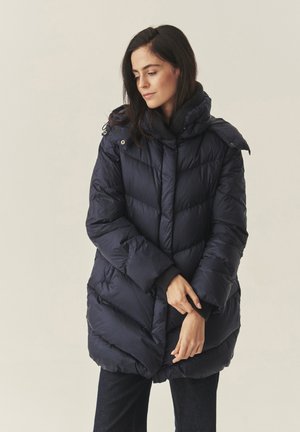 Donna con capelli scuri che indossa un cappotto imbottito navy blu con colletto alto, un maglione nero sotto e pantaloni scuri, mentre posa su uno sfondo neutro.