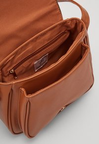 Ruskea keinonahkainen käsilaukku, jossa on pehmeä pinta, sisältää vetoketjutaskun sisäpuolella, kaksi avointa osastoa ja suorakulmainen muoto. Kultaiset metalliyksityiskohdat.