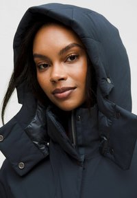 La giacca puffer blu navy presenta un cappuccio trapuntato, bottoni a scatto e una chiusura con zip. Il materiale sembra liscio con una leggera lucentezza.