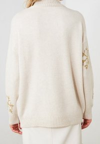 Übergroßer cremefarbener Pullover mit einem gerippten Kragen und Bündchen, der goldgestickte Schneeflockenmuster an den Ärmeln aufweist. Weicher Stoff.