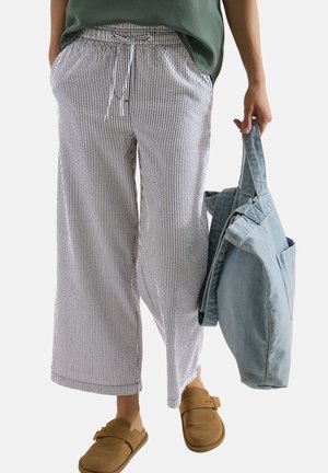 Osoba nosząca luźne, paskowane spodnie, brązowe buty wsuwane i zieloną koszulę, trzymająca w jednej ręce dużą jasnoniebieską materiałową torbę.