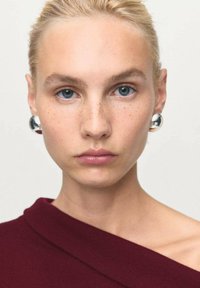 Młoda kobieta z blond włosami związanymi w kucyk, niebieskimi oczami, piegami, srebrnymi kolczykami w kształcie kół, nosząca bordową bluzkę bez ramiączek na jednolitym, jasnym tle.