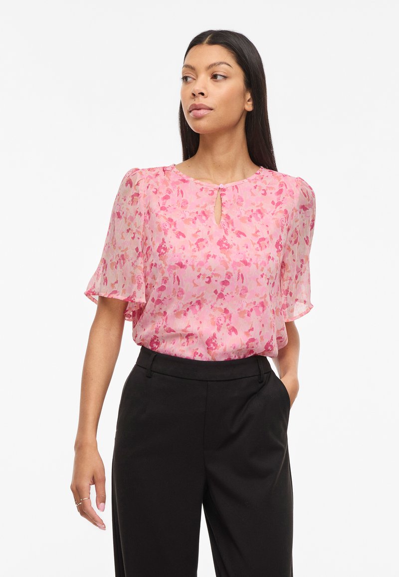 Femme portant un chemisier rose à fleurs à manches courtes avec un col en forme de trou de serrure, associé à un pantalon noir taille haute, debout avec une main dans la poche.