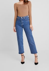 Brun långärmad bodysuit med avskuren axeldesign, kombinerad med högmidjade blå jeans i denim med raka ben och klassisk femficksstil.