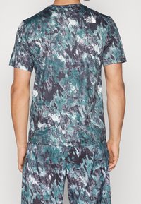 Hombre con camisa de manga corta y pantalones con un patrón de camuflaje abstracto en teal, negro y blanco, con el logo de The North Face en la parte superior de la espalda.