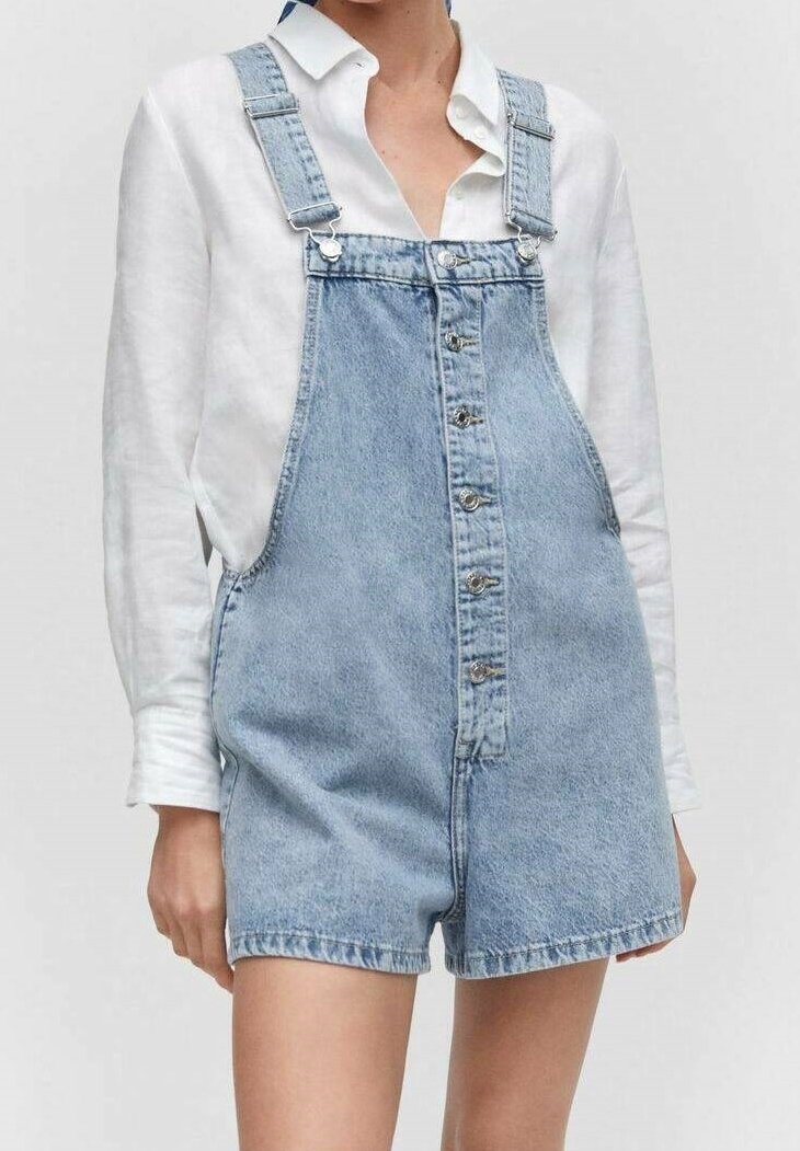 Salopette en denim bleu clair avec des jambes courtes, dotée de bretelles ajustables et d'une fermeture à boutons sur le devant. Associée à une chemise blanche à boutons.