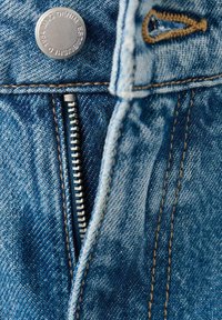 Țesătura albastră din denim include un fermoar cu dinți argintii, un nasture metalic și cusături contrastante de culoare bej de-a lungul cusăturilor.