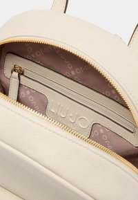 Innenseite eines cremefarbenen Leder-Rucksacks mit einem Reißverschlussfach, goldfarbigem Reißverschluss und einem rosa Futter mit Logomuster. Logo auf dem Fach geprägt.