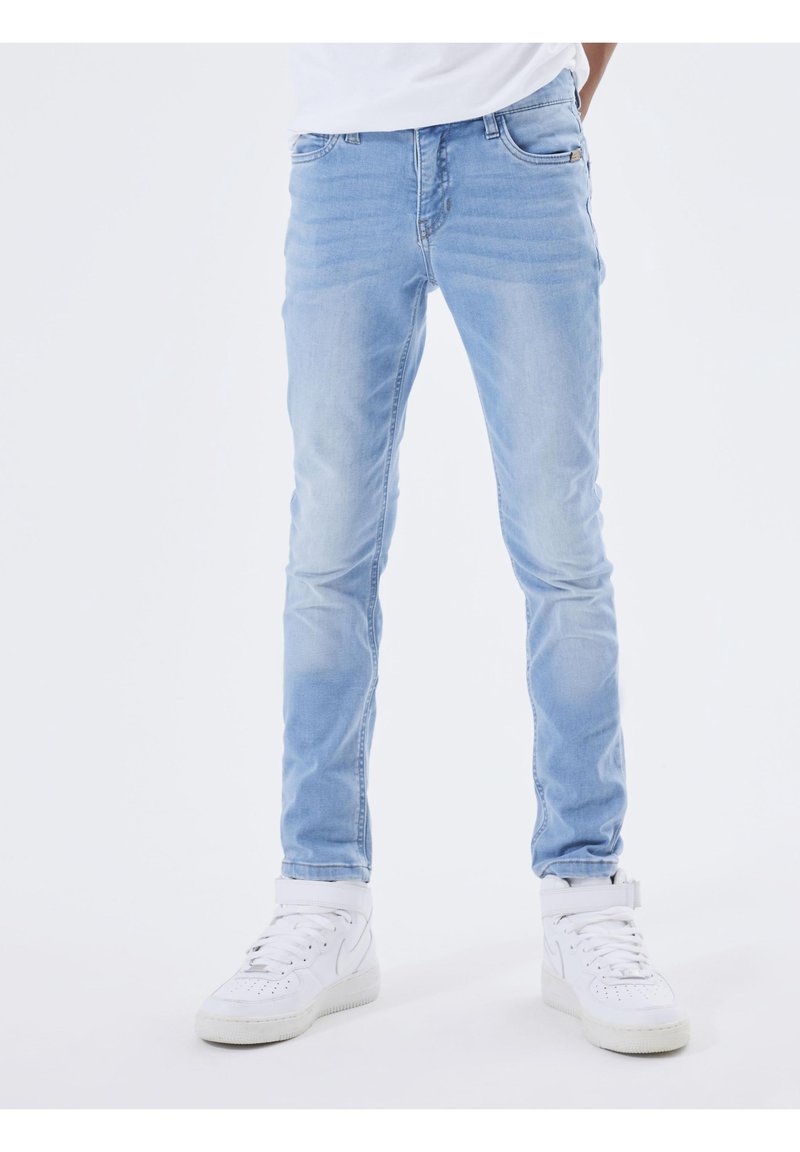Personne portant un jean slim bleu clair et des baskets montantes blanches, debout devant un fond uni.