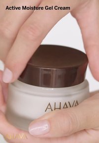 AHAVA Active Moisture Gel Cream i en rund, frostad vit behållare med ett glänsande brunt lock, hålls i en hand. Slät yta, minimal text.