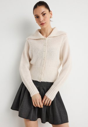 Gestrickte Strickjacke in Creme mit weitem Kragen, Knopfverschluss und geripptem Bund. Kombiniert mit einem grauen, gestreiften ausgestellten Rock.