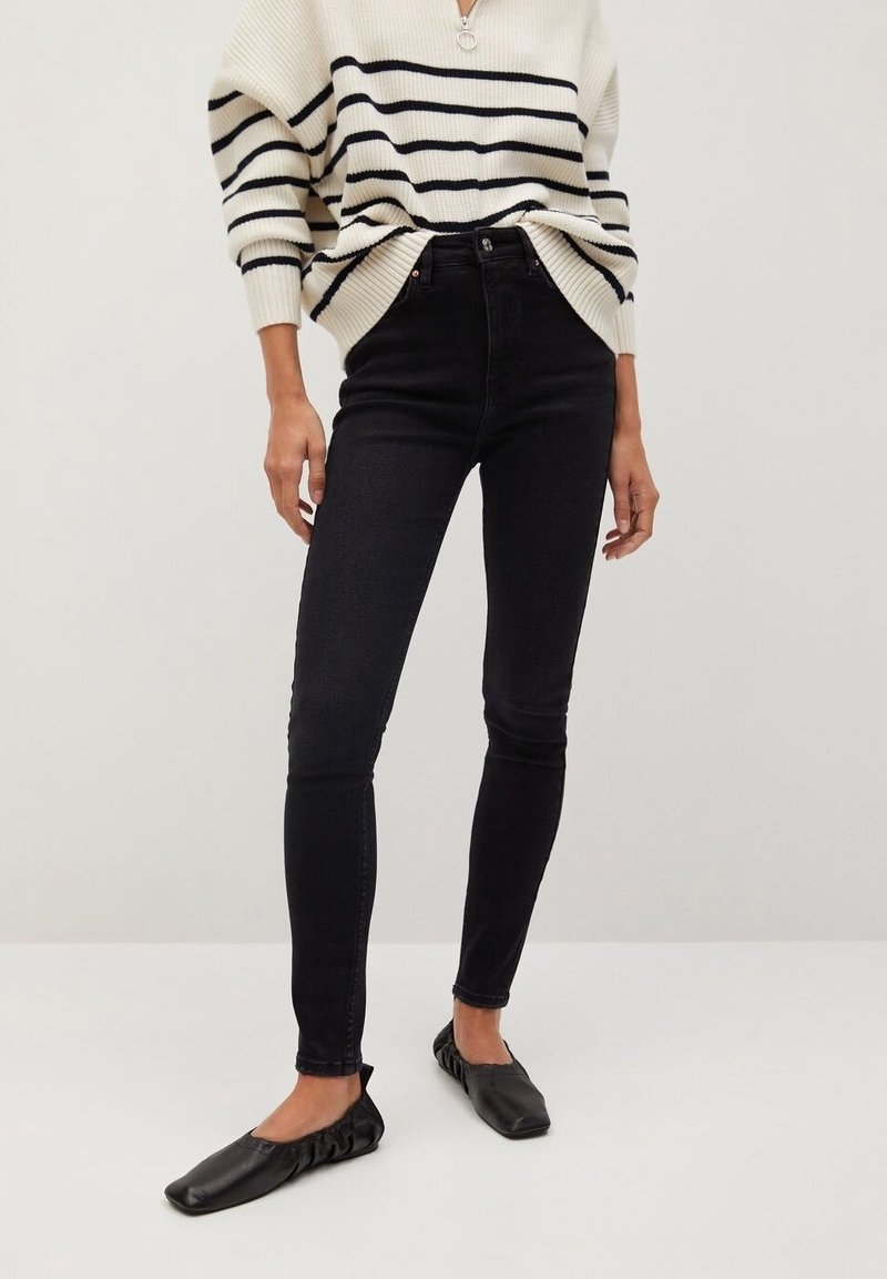 Personne portant un jean skinny noir, des chaussures plates noires et un pull blanc avec des rayures horizontales noires et une courte fermeture éclair au niveau du cou.