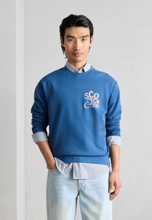 Mężczyzna w niebieskiej bluzie Scotch & Soda na paskowanej koszuli i jasnoniebieskich dżinsach, stojący z jedną ręką w kieszeni na tle jednolitej ściany.