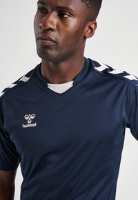 Homme portant un maillot de sport bleu marine avec des bandes en chevrons blanches sur les épaules et un logo "hummel" sur la poitrine, regardant sur le côté.