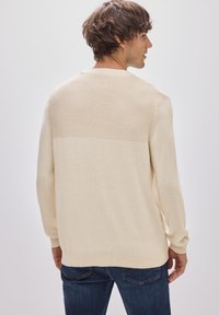 Beige stickad tröja med ribbat mönster på övre ryggen, långa ärmar och rundad halsringning, buren med mörkblå jeans.