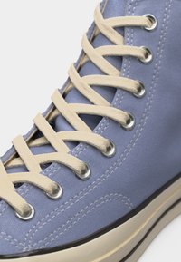 Ανοιχτού γαλάζιου καμβά αθλητικό παπούτσι με κορδόνια εκρού περασμένα από μεταλλικές τρύπες και λευκή λαστιχένια σόλα με μαύρο περίγραμμα.