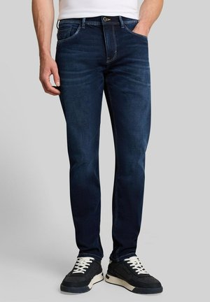 Mann trägt dunkelblaue Slim-Fit-Jeans mit weißen und schwarzen Sneakers, steht mit einer Hand in der Tasche vor schlichtem Hintergrund.