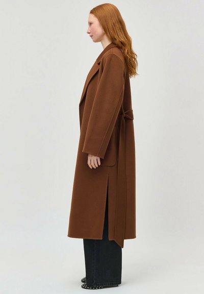 Femeie cu păr roșu, purtând o haină lungă maro, cu curea, peste pantaloni negri largi, stând în profil pe un fundal simplu.