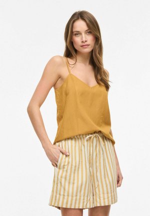 Donna che indossa una canottiera giallo senape e pantaloncini crema con righe verticali giallo senape, in piedi con una mano in tasca davanti a uno sfondo bianco.