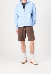 Ljusblå zip-up jacka, bruna shorts och krämfärgade sneakers med röda ränder. Grå strumpor synliga. Avslappnad, luftig passform.