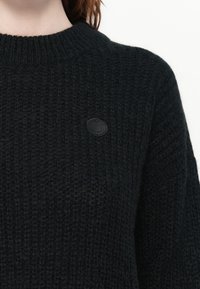 Zwart gebreid trui met een geribbelde textuur, voorzien van een ronde halslijn en een klein reliëflogo op de voorkant.