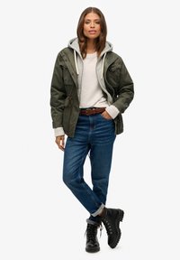 Chaqueta verde de estilo militar sobre una sudadera gris, combinada con un suéter claro y jeans azules. Botas negras de cordones completan el atuendo.