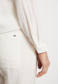 Chemise blanche texturée à manches longues. Poignet à revers montrant une fermeture à bouton. En dessous, tissu de couleur claire avec une poche plaquée et un détail de logo.