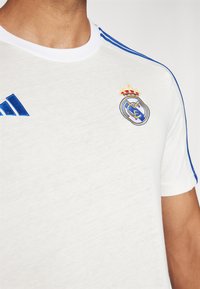Bijela pamučna majica s plavim Adidas trakama na ramenima. Izvezeni okrugli logo s krunom na lijevoj strani prsnog dijela.