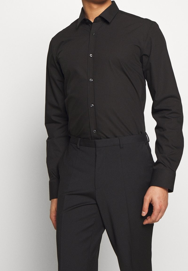 Homme portant une chemise cintrée noire à manches longues boutonnée, rentrée dans un pantalon habillé noir, devant un fond uni.
