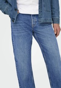 Jeans de mezclilla azul presentan un diseño de pierna recta, lavado claro, un ligero desgasto y costuras clásicas. Combinados con una chaqueta de mezclilla y una camiseta blanca.