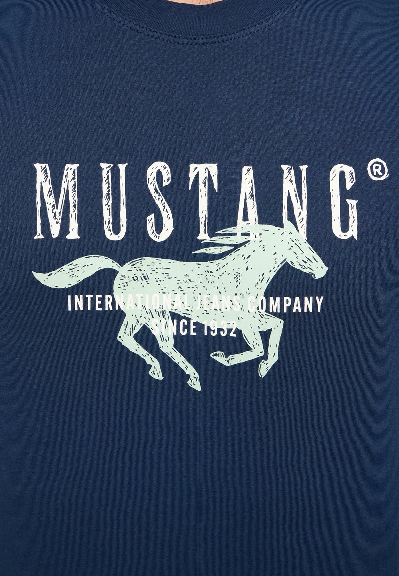 T-shirt en coton bleu marine avec un graphique de cheval au galop vert clair et un texte blanc disant "MUSTANG INTERNATIONAL JEANS COMPANY SINCE 1952."