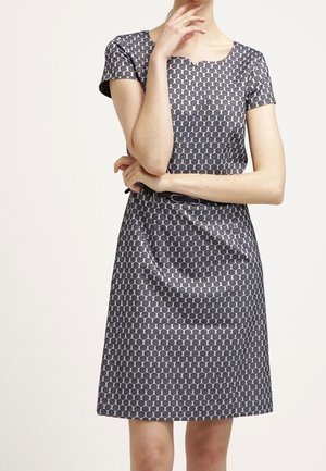 Femme portant une robe à manches courtes, longueur genoux, avec un motif circulaire noir et gris et une fine ceinture noire, debout les bras croisés.