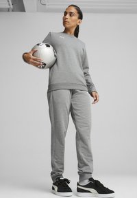 Felpa grigia in pile e pantaloni della tuta abbinati, entrambi con un design semplice. Sneakers nere con dettagli bianchi. Tiene un pallone da calcio.