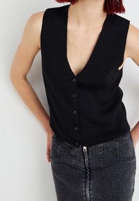 Svart ärmlös stickad topp med djup V-ringning, prydd med sex framknappar och ribbat mönster. Matchas med en högmidjad grå jeanskjol.