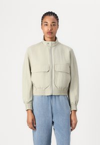 Veste courte grise clair avec un col haut, des poignets élastiques et deux poches avant. Le tissu a un aspect texturé.