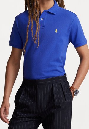 Polo bleu à manches courtes avec logo brodé jaune, porté avec un pantalon noir à rayures fines et une montre en cuir marron au poignet gauche.