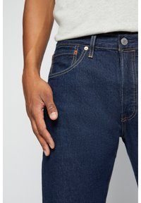 Hombre que lleva jeans de mezclilla azul oscuro y una camisa gris clara, con la mano descansando cerca del bolsillo frontal derecho de los jeans.